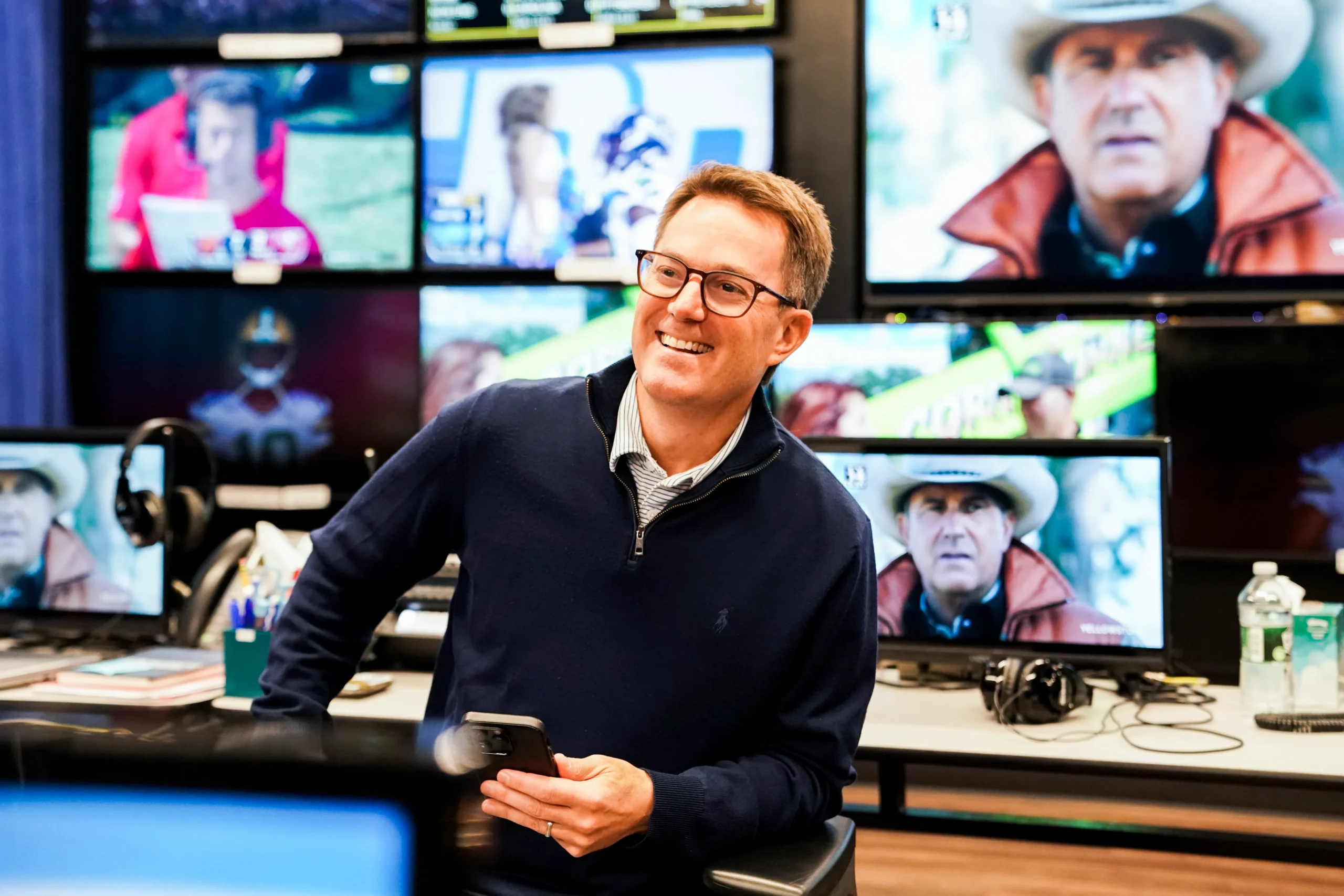 A smiling man with glasses holds a phone in a control room filled with television monitors.