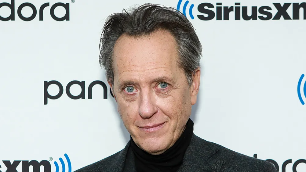 A man with gray hair and blue eyes poses in a dark blazer before SiriusXM and Pandora logos.