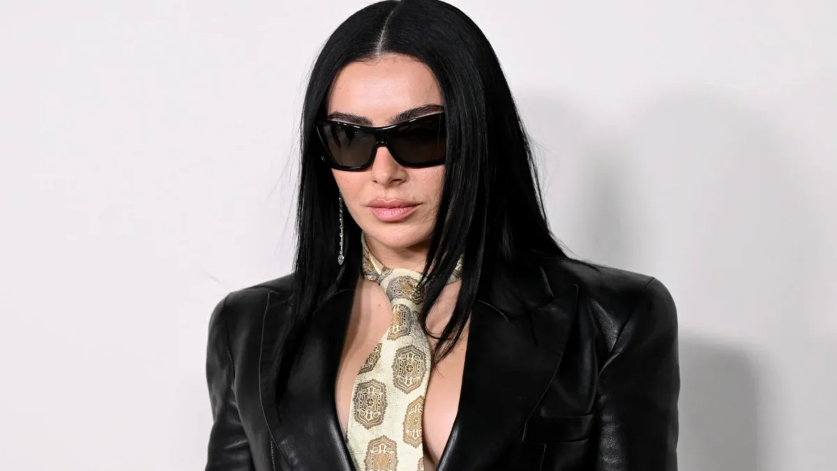 A woman with long black hair wears oversized sunglasses, a black leather jacket, and a patterned scarf.