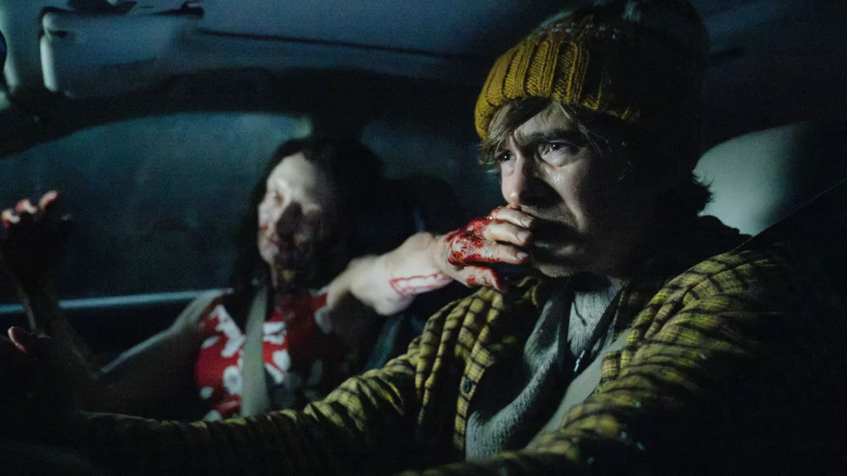 A person in a yellow beanie grips the wheel as a bloodied hand covers their mouth in a dark car.