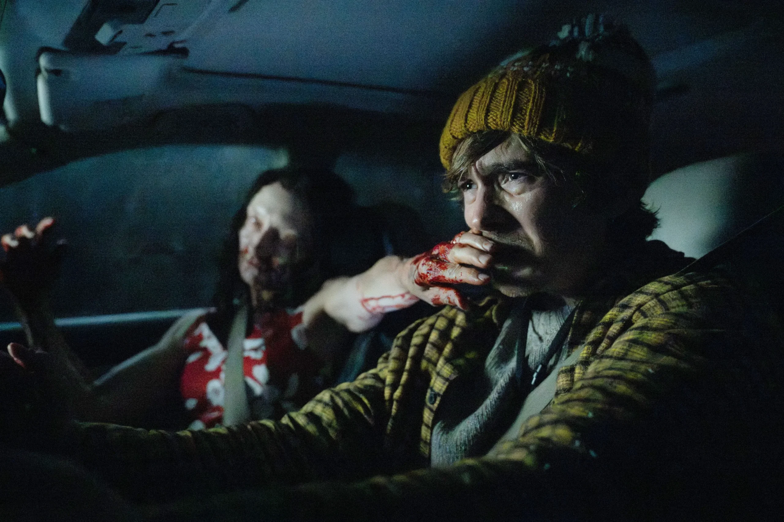 A person in a yellow beanie grips the wheel as a bloodied hand covers their mouth in a dark car.