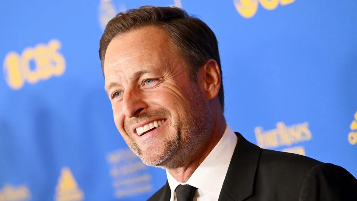 Smiling man in a black suit and tie poses against a blue backdrop with blurred yellow logos.