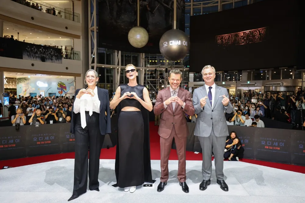 Four people pose on a red carpet before a large crowd inside a multi-level venue.