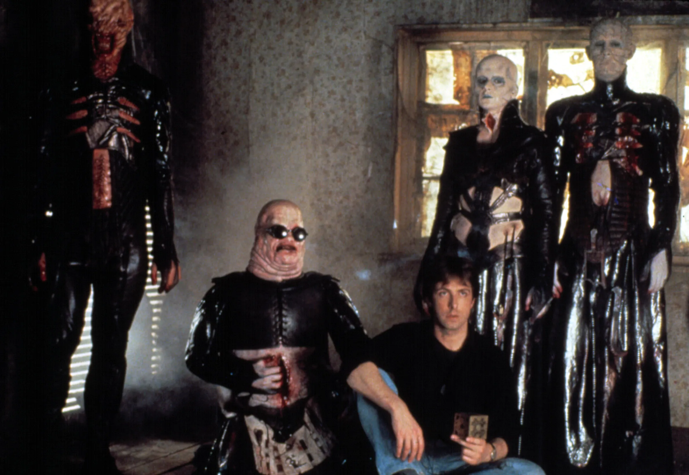 Five grotesque figures in glossy black costumes surround a seated man in a dim, decaying room.