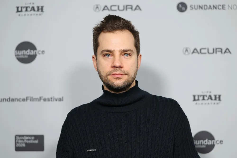 A man in a black turtleneck sweater poses before a gray backdrop with festival and sponsor logos.