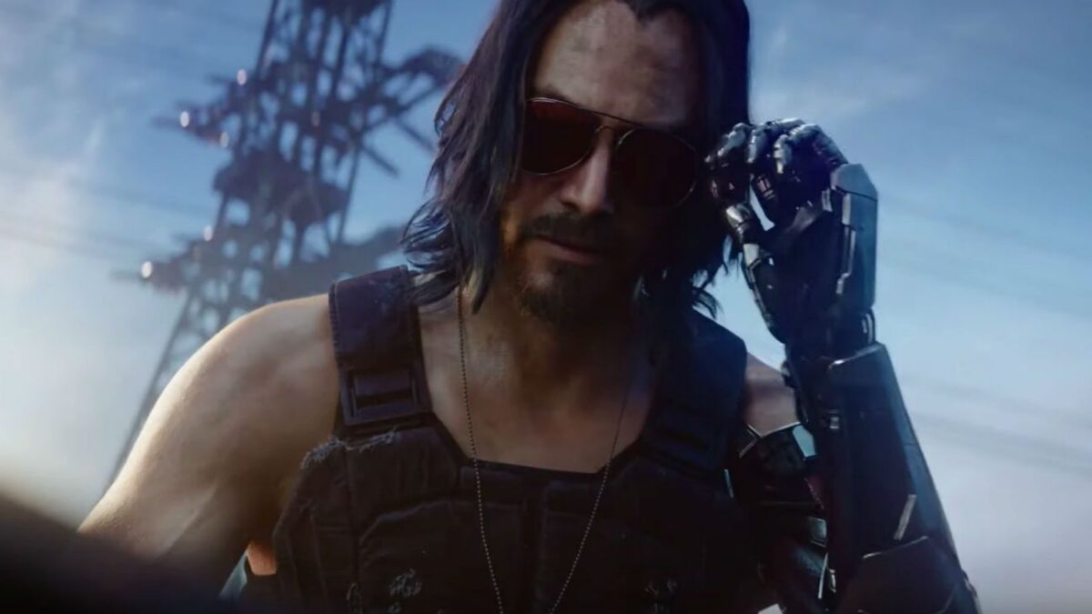 A long-haired man in sunglasses touches his temple with a robotic hand before an industrial tower.