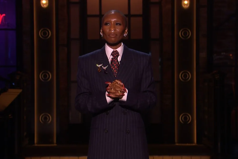 A person in a pinstripe suit stands on a dimly lit stage with hands clasped.