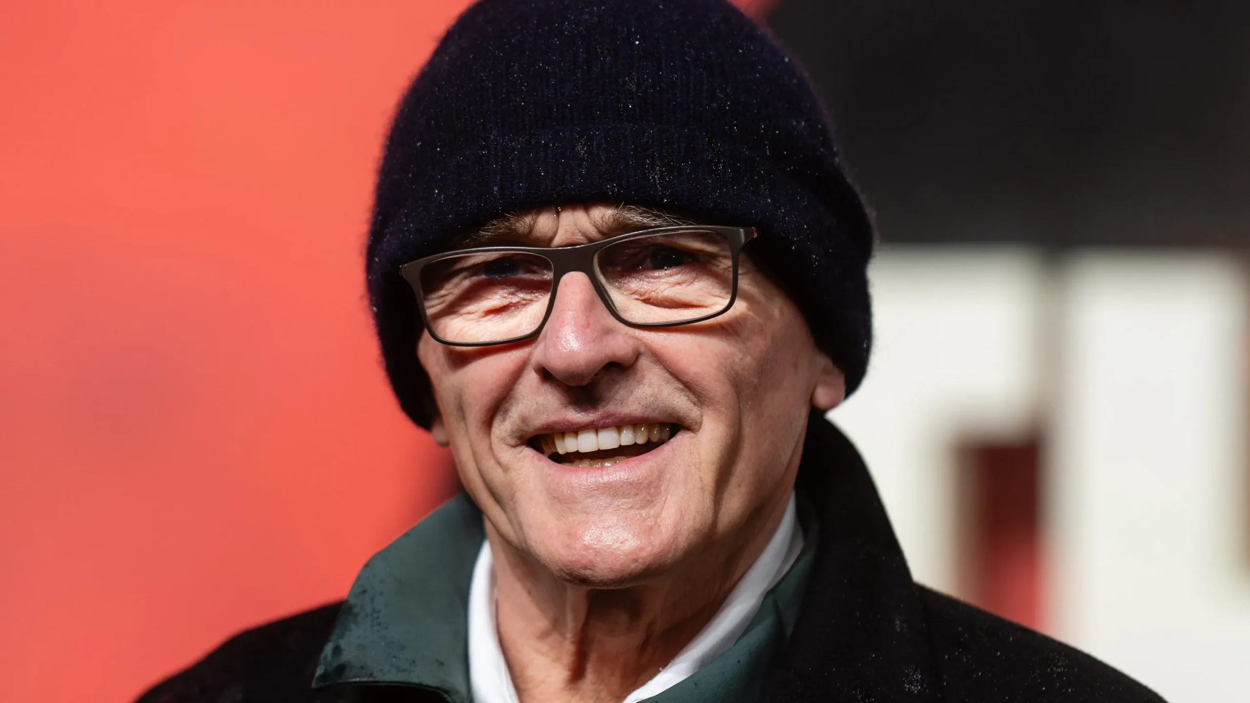 A smiling older man wearing glasses and a black knit beanie stands before a red background.