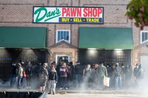 A crowd stands outside a brick pawn shop storefront as smoke drifts across the street.