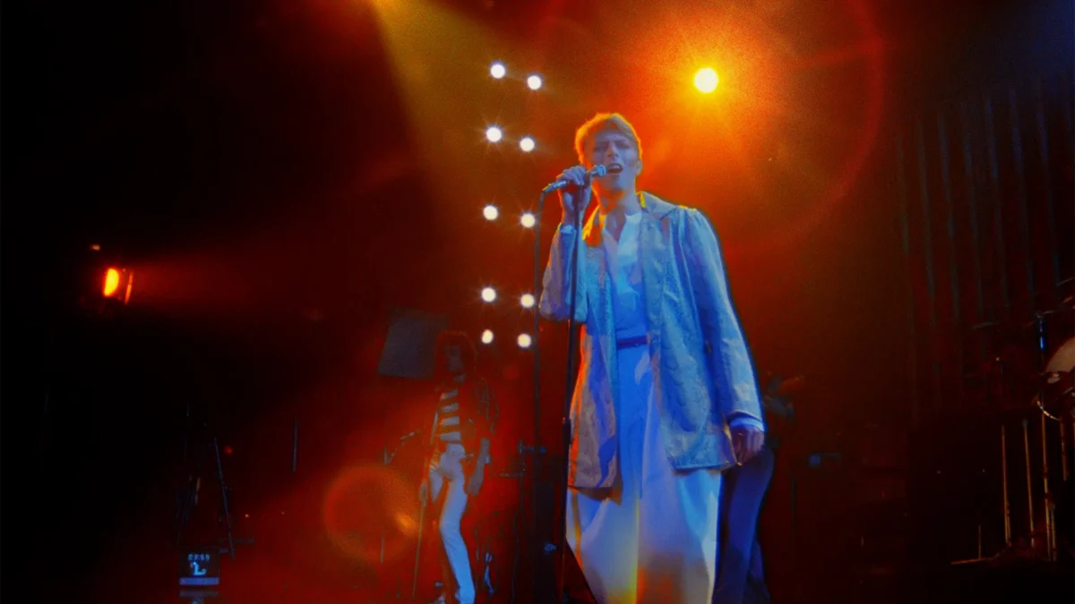 A singer in a light suit performs at a microphone under red and orange stage lights.
