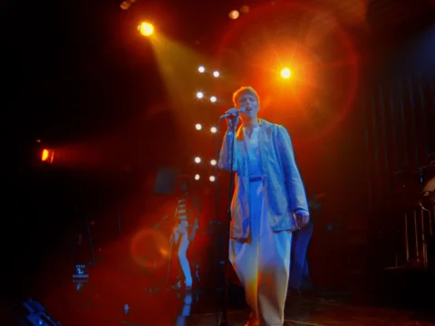 A singer in a light suit performs at a microphone under red and orange stage lights.