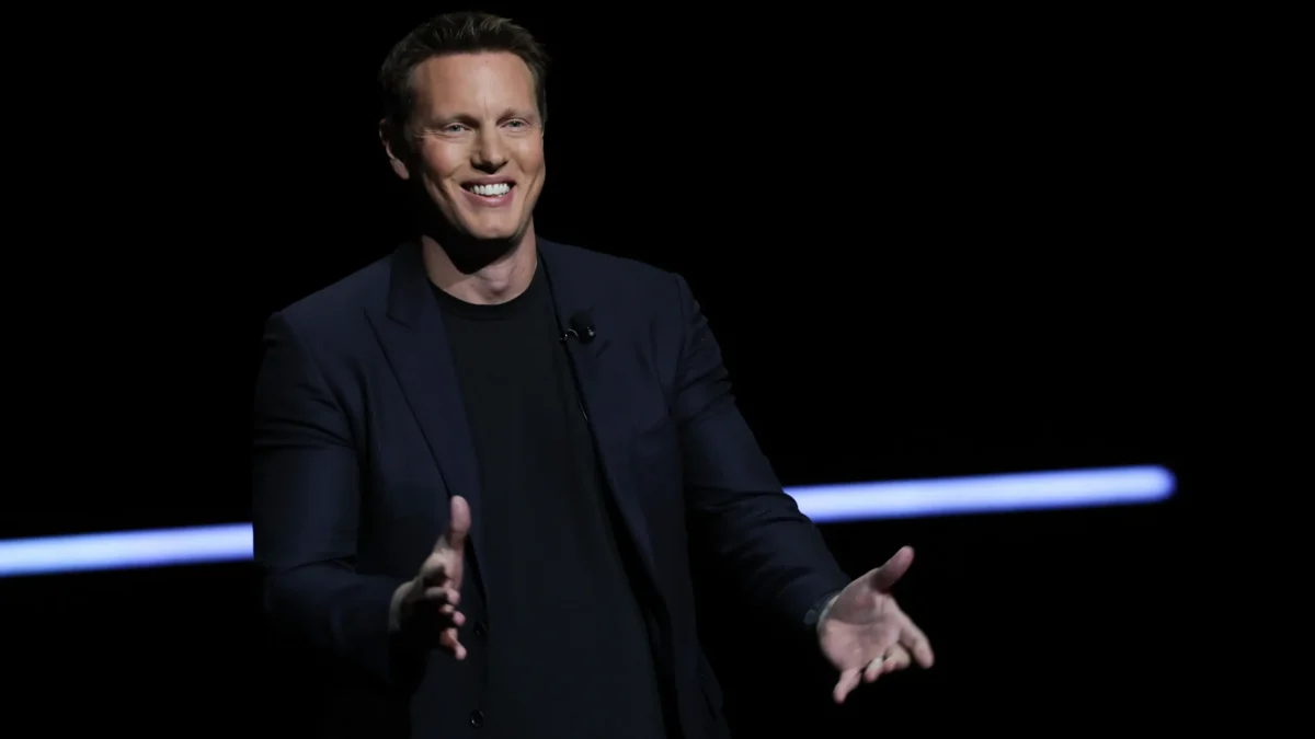 A smiling man in a dark blazer gestures with both hands against a black background.