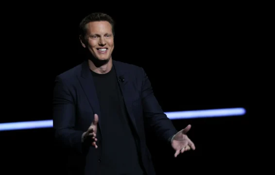 A smiling man in a dark blazer gestures with both hands against a black background.