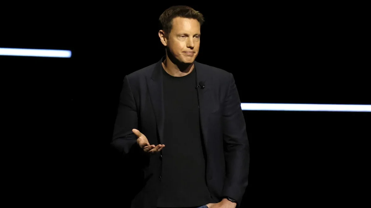 A man in a dark blazer and jeans gestures on a dimly lit stage with horizontal light bars.