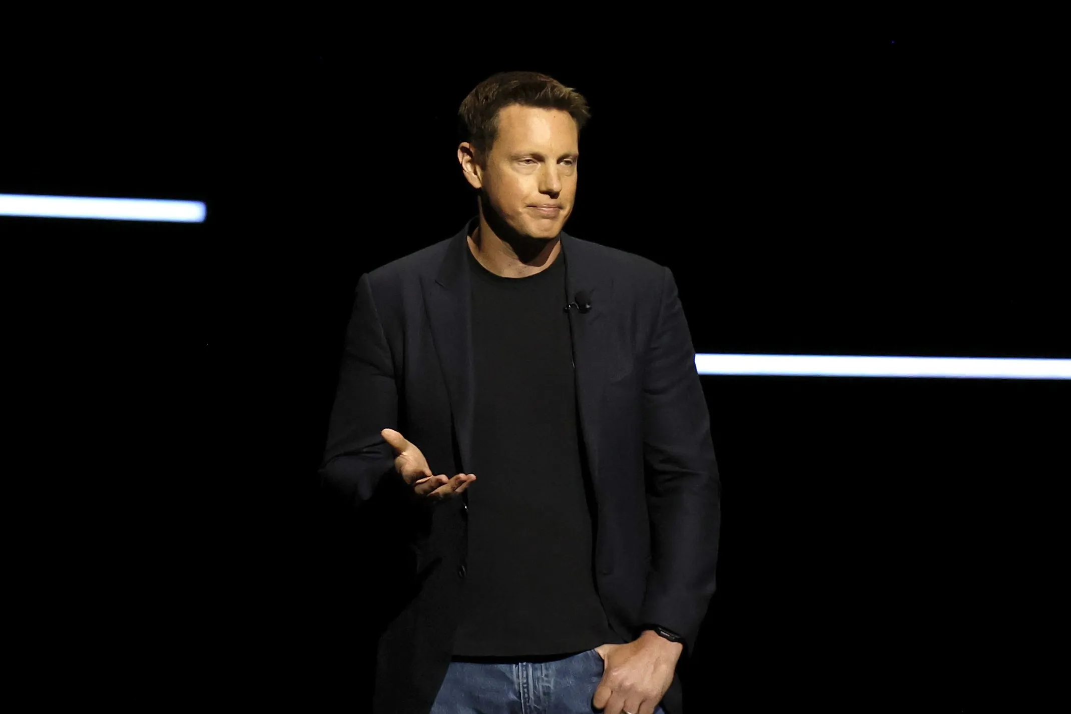 A man in a dark blazer and jeans gestures on a dimly lit stage with horizontal light bars.