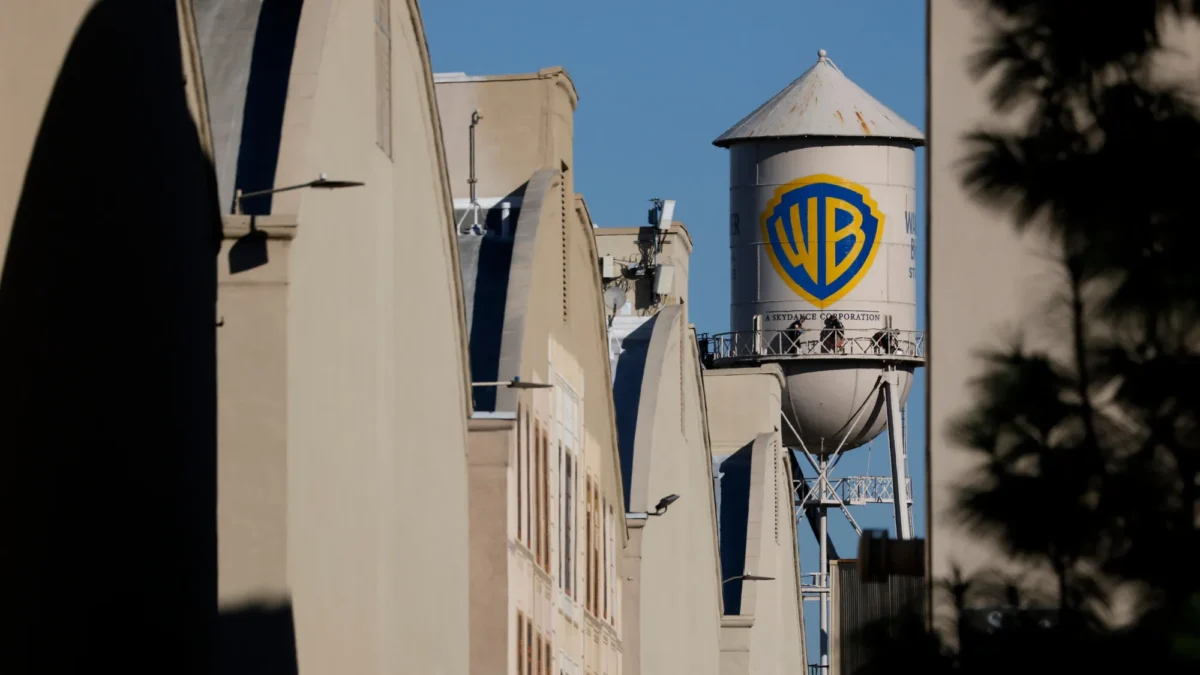 A water tower with a WB logo rises behind beige buildings under a clear blue sky.