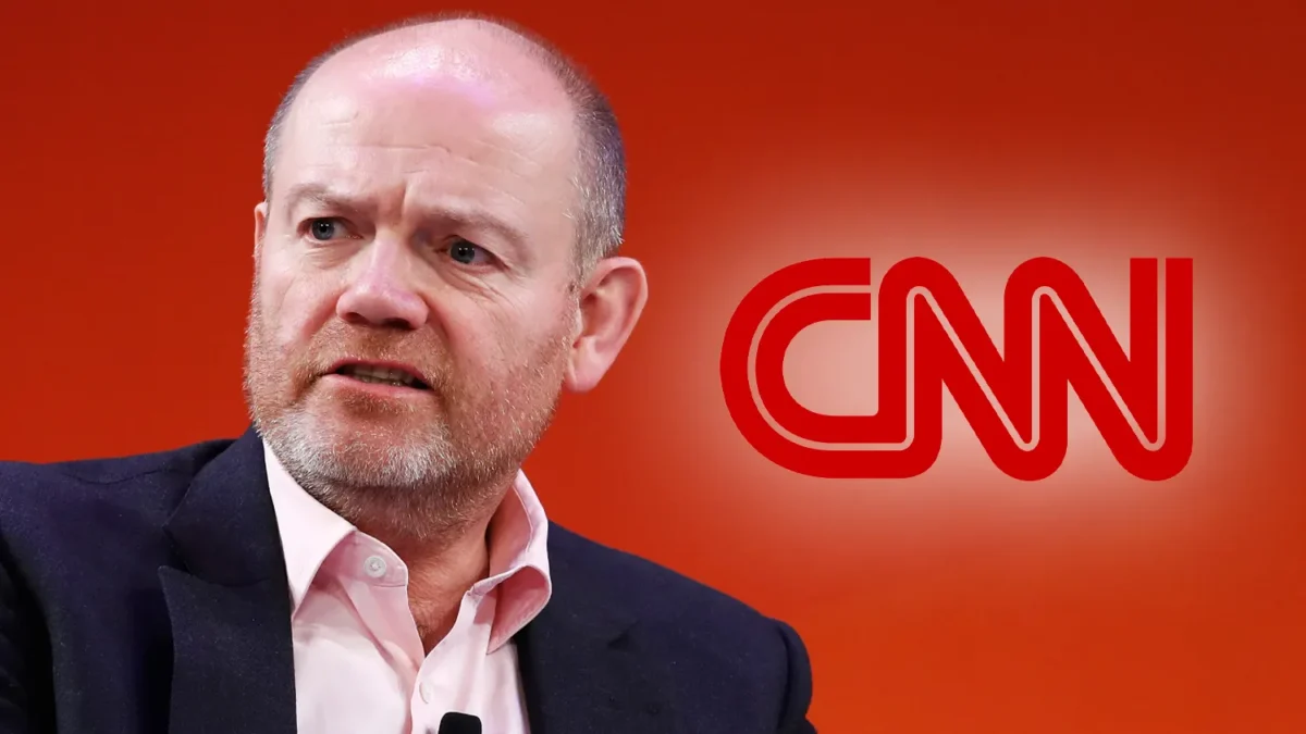 Balding man in a dark blazer sits before a red background with a large CNN logo.
