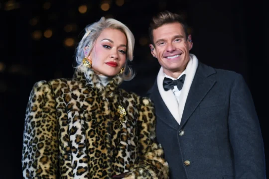 Two people pose against a dark background, one in a leopard-print coat and one in a gray coat.
