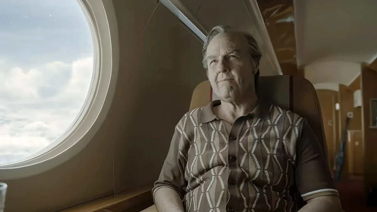 An older man sits beside an airplane window, looking toward the bright clouds outside.