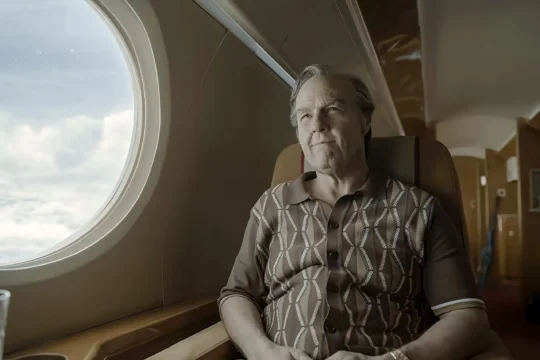An older man sits beside an airplane window, looking toward the bright clouds outside.