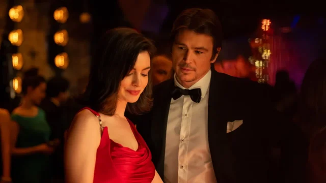 A woman in a red dress and a man in a tuxedo stand together at a dimly lit formal gathering.