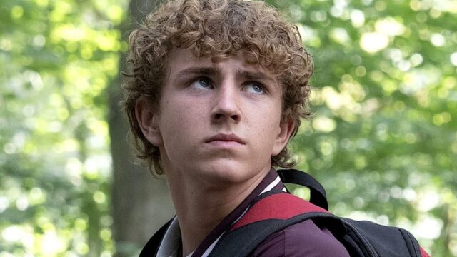 A curly-haired teenager with a backpack looks off to the side in a sunlit forest.