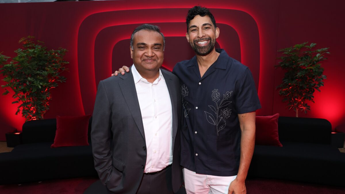 Two smiling men pose together in front of a red illuminated backdrop with plants and lounge seating.
