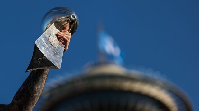 A tattooed arm holds a silver football-shaped trophy against a blue sky.