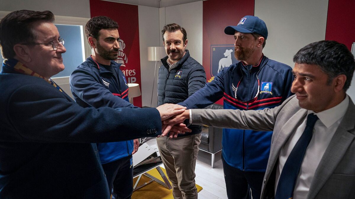 Five men stand in a room with their hands stacked together, wearing suits and matching athletic jackets.