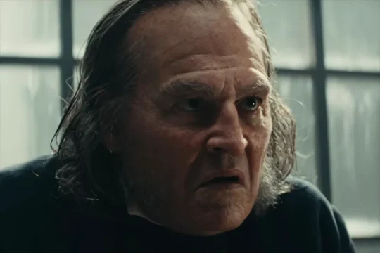 An older man with long gray hair frowns in a dim room before a window.