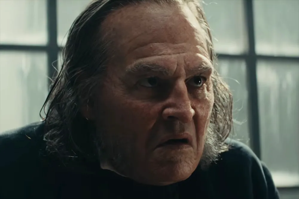An older man with long gray hair frowns in a dim room before a window.