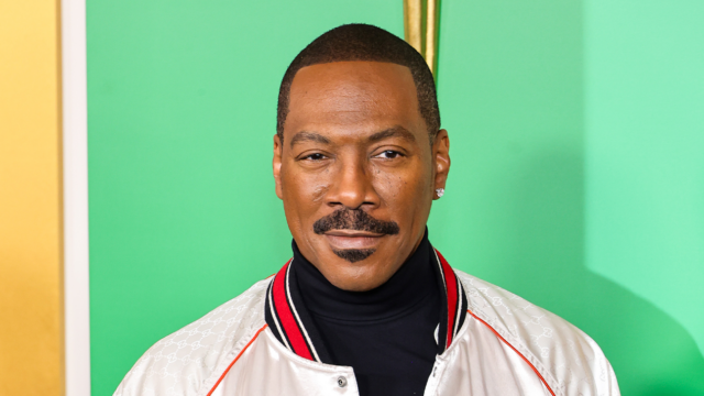 A man with a mustache wears a white jacket over a black turtleneck against a green backdrop.