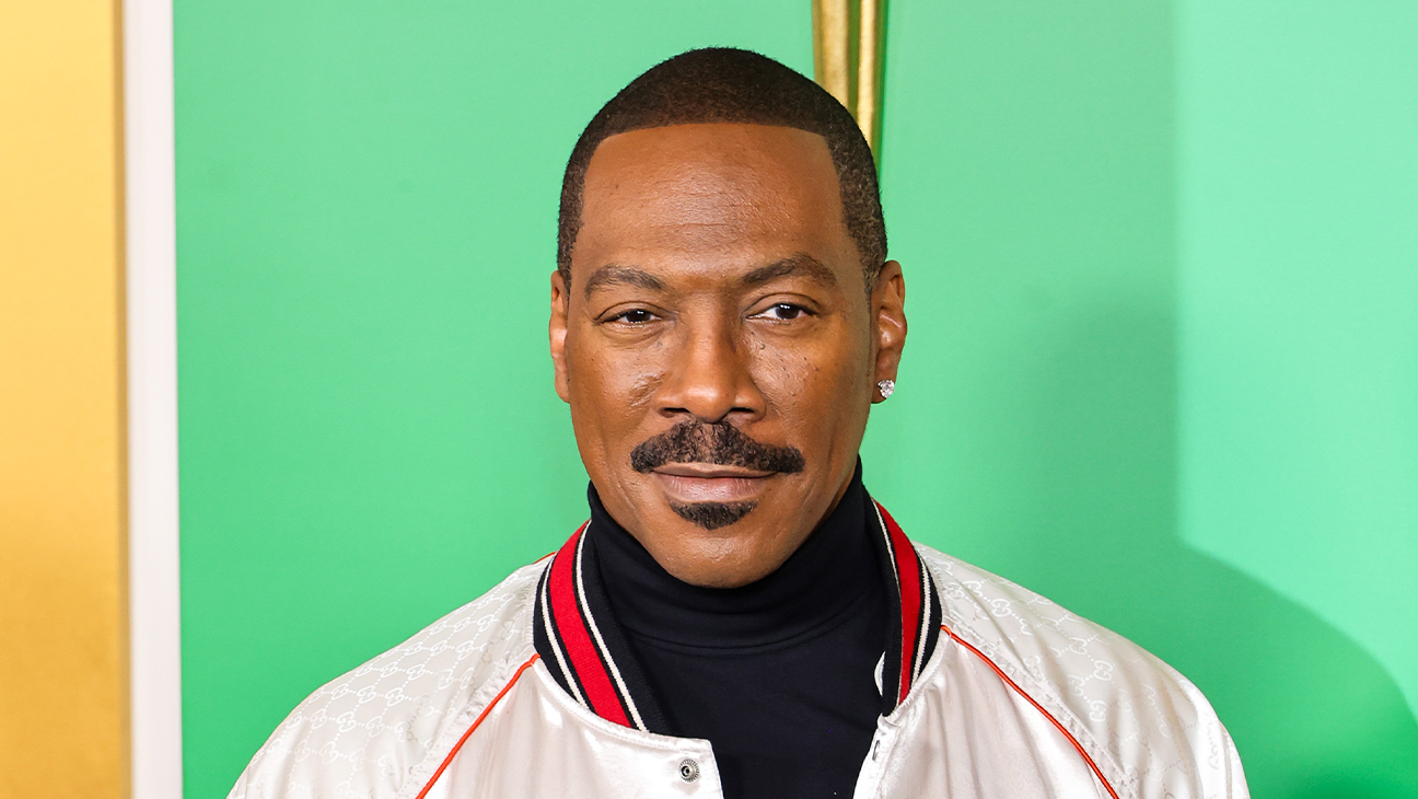 A man with a mustache wears a white jacket over a black turtleneck against a green backdrop.