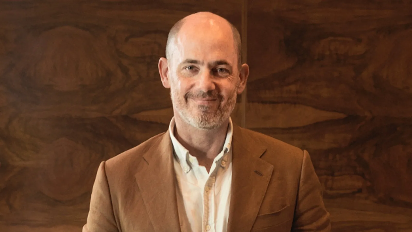 A smiling bald man in a brown blazer stands before a dark wood-paneled wall.