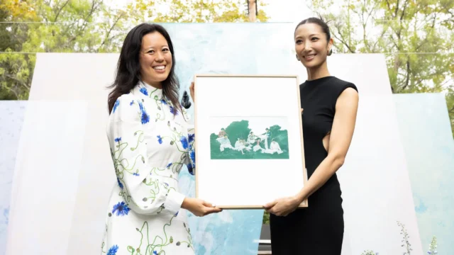 Two smiling women hold a framed artwork outdoors in front of pale blue and white panels.
