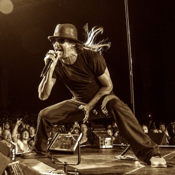 A performer in a hat sings into a microphone onstage before a cheering crowd.