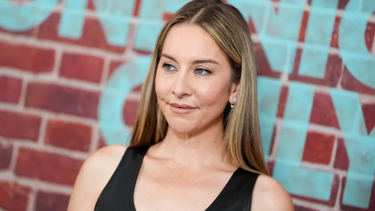 Woman with long blonde hair in a black sleeveless top poses before a red brick wall with turquoise lettering.