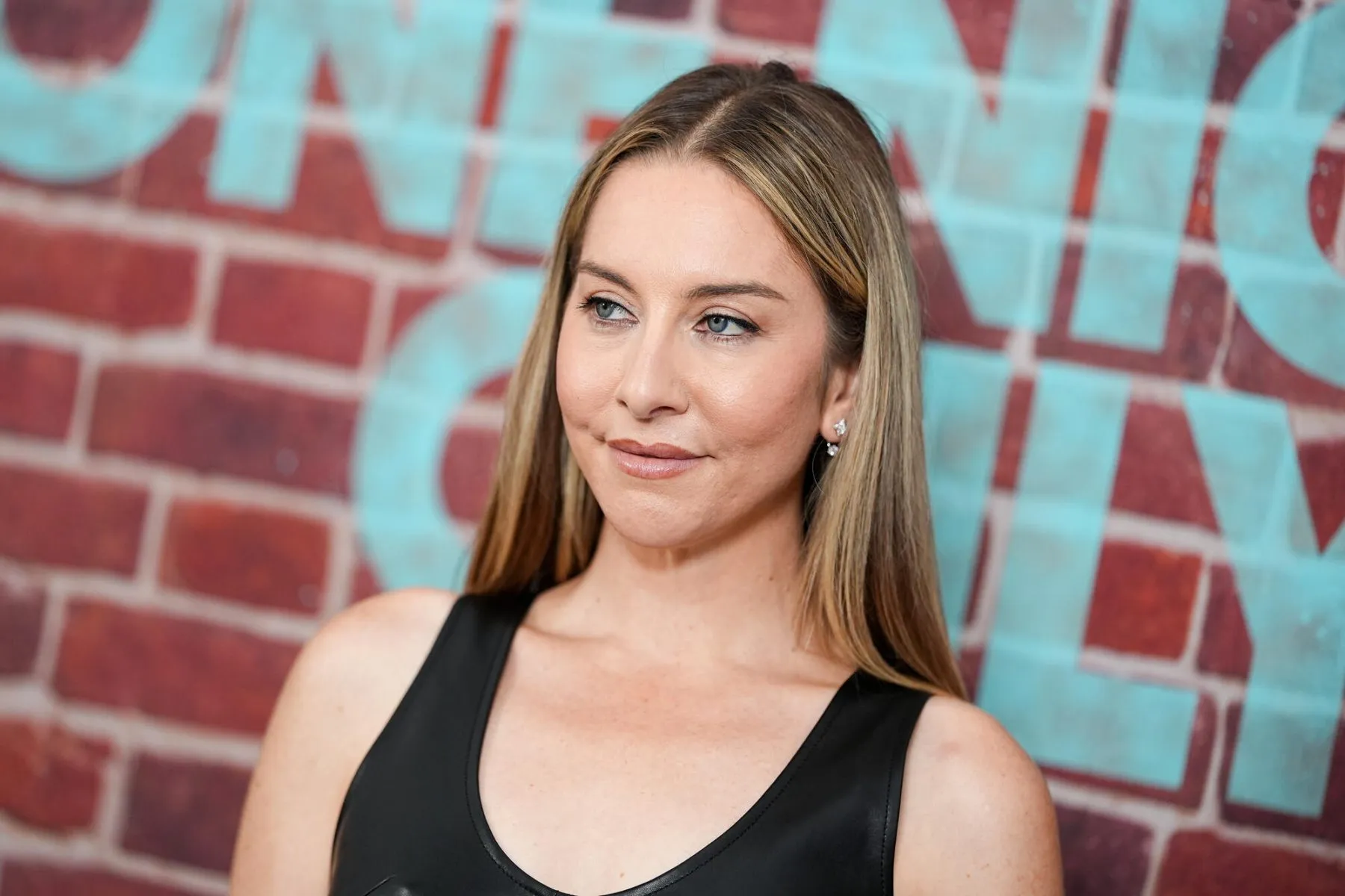 Woman with long blonde hair in a black sleeveless top poses before a red brick wall with turquoise lettering.