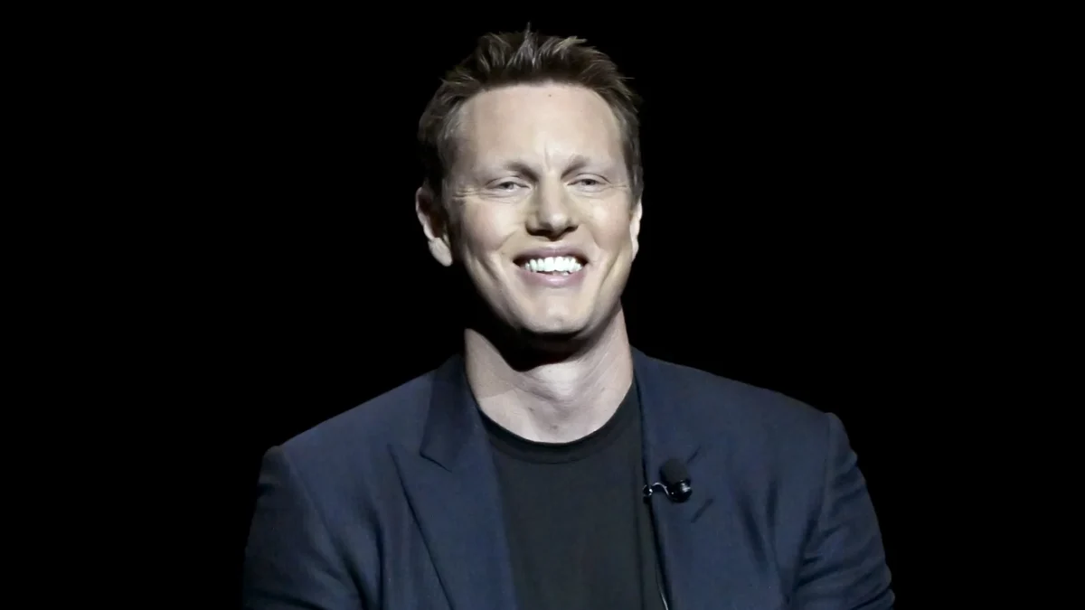 A smiling man in a dark blazer and black shirt sits against a black background.