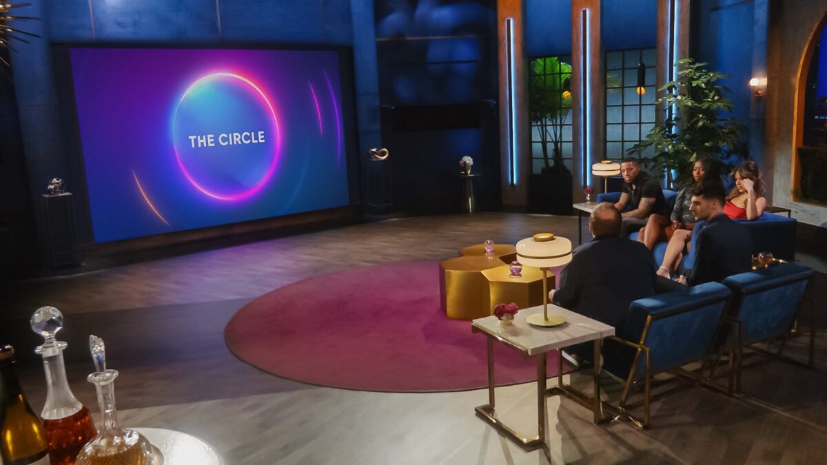 A group sits in a lounge facing a large screen displaying “The Circle” with colorful neon rings.