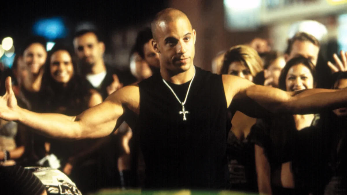 A bald man in a sleeveless black shirt spreads his arms before a cheering nighttime crowd.