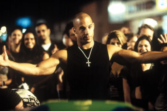 A bald man in a sleeveless black shirt spreads his arms before a cheering nighttime crowd.