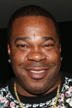 Close-up of a smiling man wearing stud earrings, a gold chain, and a floral-patterned shirt.