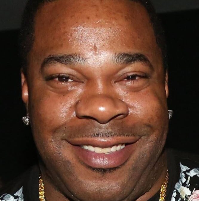 Close-up of a smiling man wearing stud earrings, a gold chain, and a floral-patterned shirt.