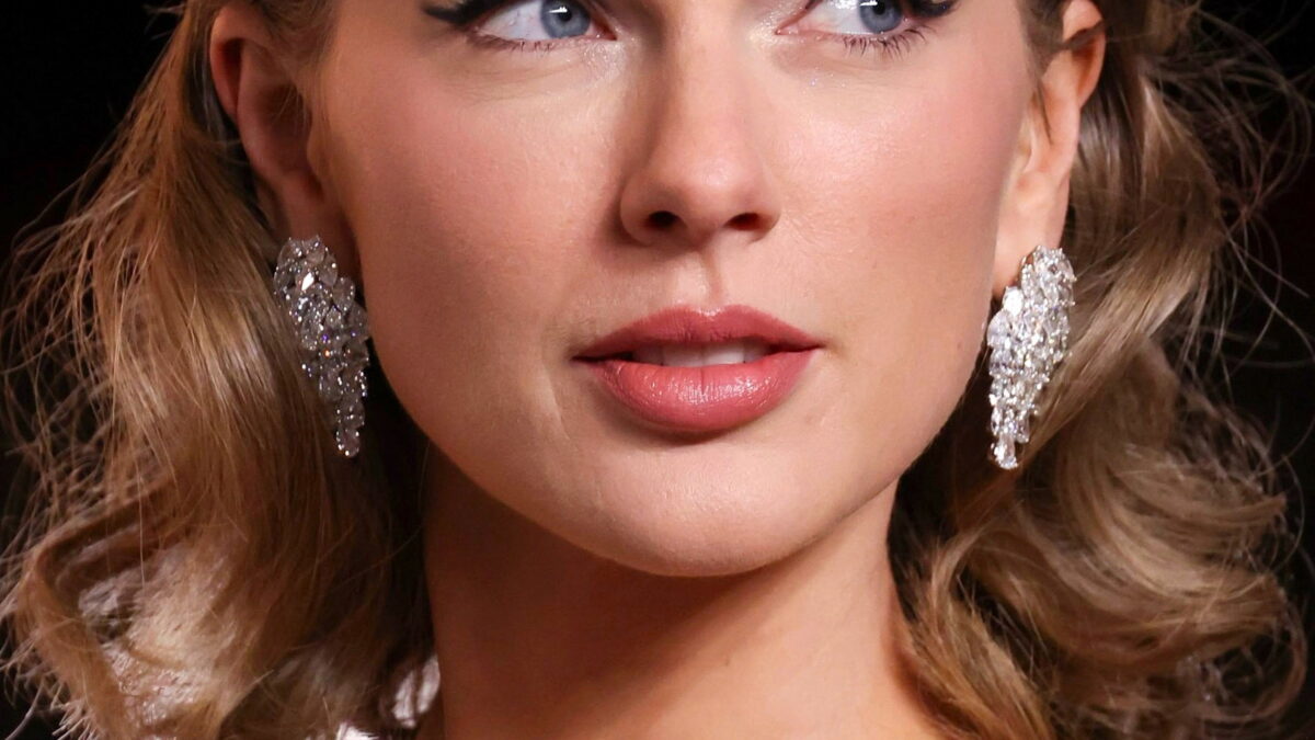 Close-up portrait of a woman with wavy blonde hair, jeweled earrings, and red lipstick looking upward.