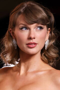 Close-up portrait of a woman with wavy blonde hair, jeweled earrings, and red lipstick looking upward.