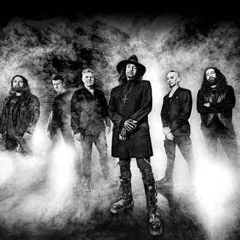 Six dark-clad people pose in a smoky black-and-white scene with the central figure wearing a wide-brim hat.