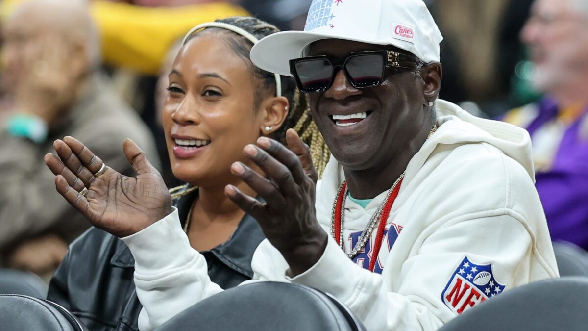 A man in sunglasses and a white cap claps while seated beside a smiling woman in a crowd.