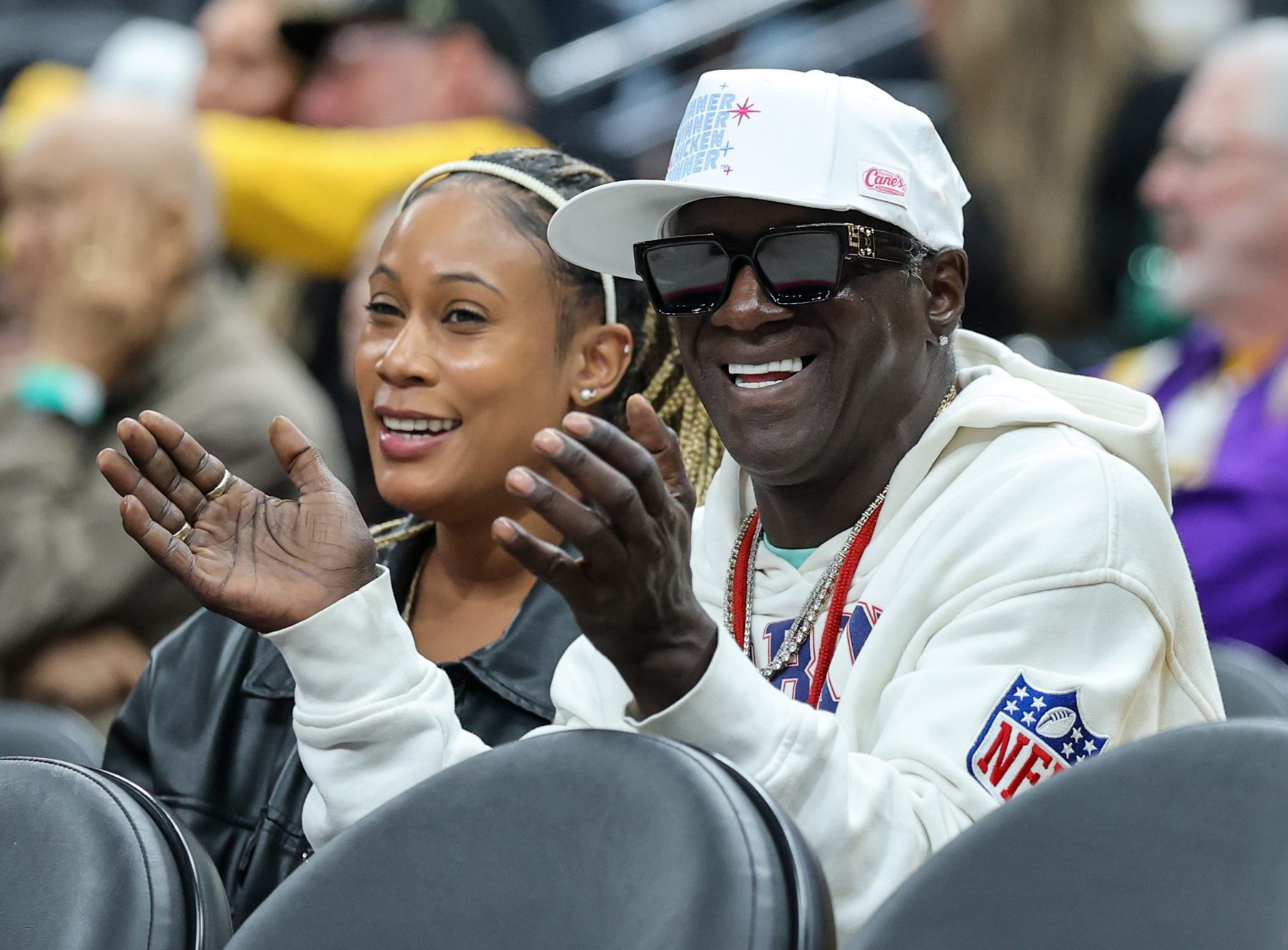 A man in sunglasses and a white cap claps while seated beside a smiling woman in a crowd.