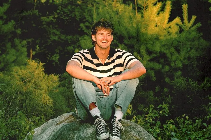 Smiling man in a striped shirt sits on a rock surrounded by green trees.
